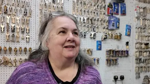 Gislea has shoulder length grey-black hair and glasses resting on her forehead. She is wearing a purple jumper and behind her is a wall of keys in her workplace. 