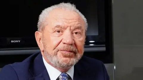 Lord Alan Sugar in a suit and tie looking pensive.