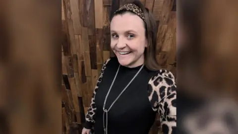 Tonia Dunn pictured in a selfie. She is smiling and stood against a wooden panel wall where she wears a leopard print sleeve shirt and silver long necklace.