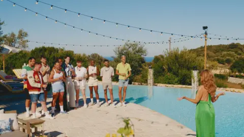 BBC/TwoFour Dannii Minogue stands at a poolside facing ten male contestants of I Kissed a Boy. She wears a green dress and has long brown hair and the boys wear t-shirts and shorts