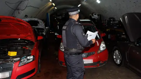 A police officer in uniform, with his back to the camera, examines a number of cars including a red Honda stashed in a lock-up with an arch-shaped ceiling. 