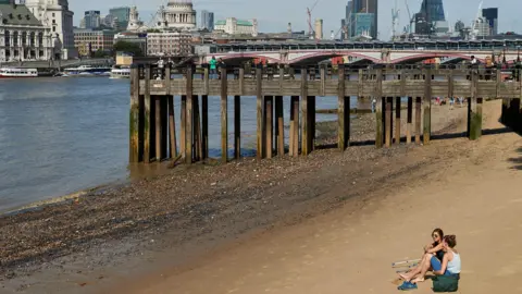Two young women pictures sitting on a beach by the river Thames, with the London skyline in the background