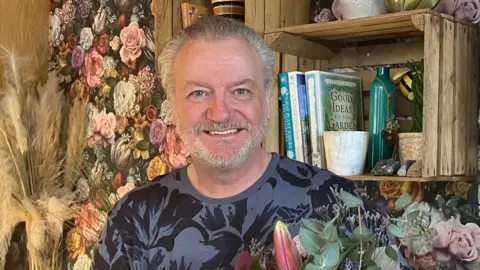 A man with short slicked back grey hair and beard stands in front of a wall of flowers