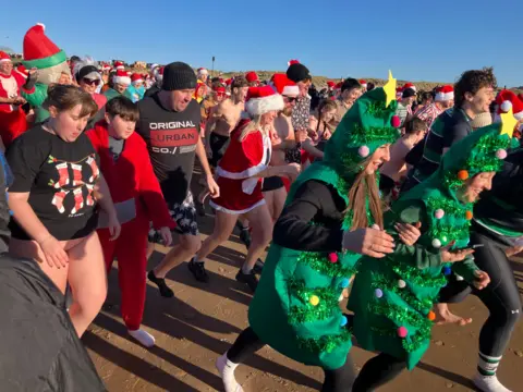 Close up of crowds running into the water. There are two ladies in front dressed up as Christmas trees. Other people in the background are wearing Christmas themed outfits with their swimming costumes. 