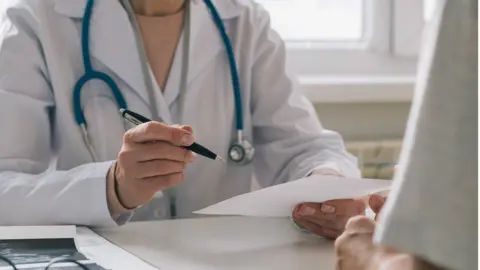 Getty Images An anonymous doctor wearing a white doctor's coat with a stethoscope around their neck while they sit at a desk with a pen in one hand and a piece of paper in the other.