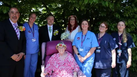 St Elizabeth Hospice A group of men and women - some nurse outfits and others in smart wedding attire - standing behind a woman with a colourful dress on who is sitting on a large chair. They are all looking into the camera and standing under some leaves.