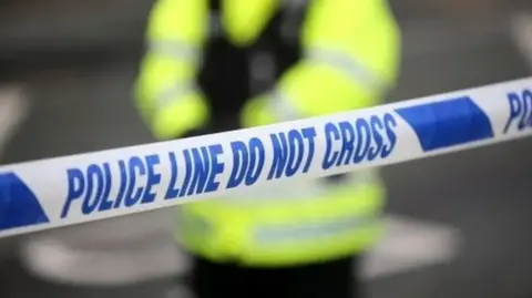 A police officer in a yellow hi-vis jacket stands behind some blue and white police tape.