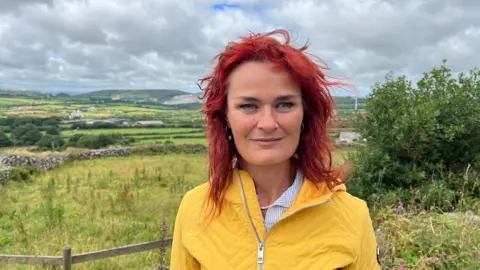 Dr Joanie Willett wearing a shirt underneath a yellow zipped jacket and standing in front of fields overlooking St Dennis in Cornwall