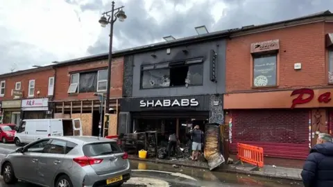 A row of restaurants, showing a restaurant called SHABABS with a smashed glass window on the first floor. The restaurant, painted black, is charred from the fire, with 2 people clearing up the mess at the front. 
