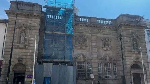 Alex Pope/BBC A wide view of a town-centre library building with scaffolding and blue netting covering part of the façade during restoration work.
The full frontage of a historic public library shows ongoing repairs, with metal hoarding at street level and scaffolding above.
Cars and pedestrians pass in front of a stone library building undergoing refurbishment, partially covered in construction materials.