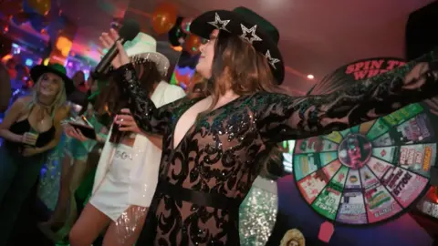 Haus of Fun A person wearing a black sparkly bodysuit and a black cowboy hat holds a microphone and dances in front of a 'spin-the-wheel' wheel. They have long brown hair.
