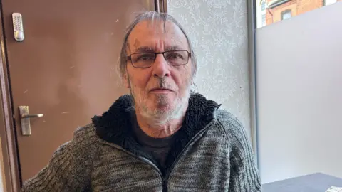 Kate Bradbrook/BBC A man wearing a grey jacket with a black top underneath. He has glasses and is looking at the camera 