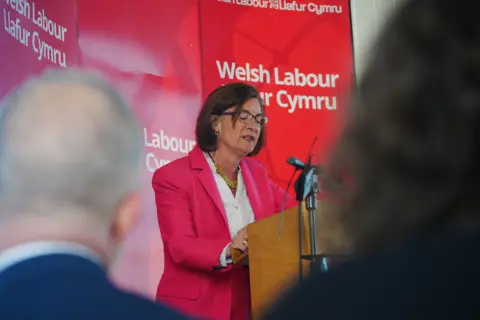 PA Media Eluned Morgan stands at a podium giving a speech. She wears a bright pink suit jacket and a white shirt. A red board stands behind her with the words Welsh Labour on. People sit and listen to her speaking, the backs of their heads are blurred.