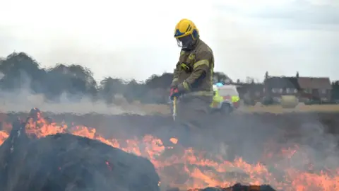 Norfolk Fire and Rescue Service A firefighter in full uniform stands in a field on fire.
