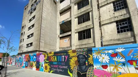 Steve Martin/BBC A derelict building which was a former silo on Ipswich waterfront. There are construction boards put up with art work which is colourful with flowers. 
