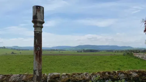 A metal post which was once a fingerpost pokes into the air next to a stone wall in front of large grassy field. Behind there is a range of low lying, rolling hills.