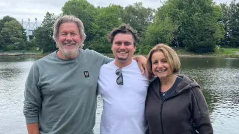Rii Schroer Alex Warwick with his parents pose arm-in-arm in front of a lake