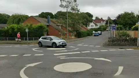 A double mini roundabout on a suburban street. Houses and parked cars can be seen. A grey car is crossing the roundabout.