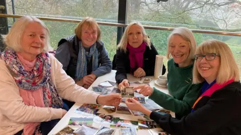 Five women with white hair sit around a table smiling. On the table is photographs from their days out together.