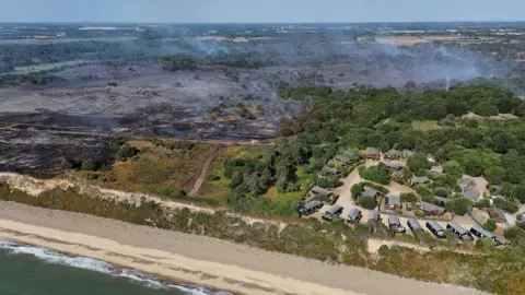 Reuters A drone image of a coastline. There is a holiday park with static caravans. The land around the site has been scorched black from a wildfire. Smoke billows into the air from some of the land. 