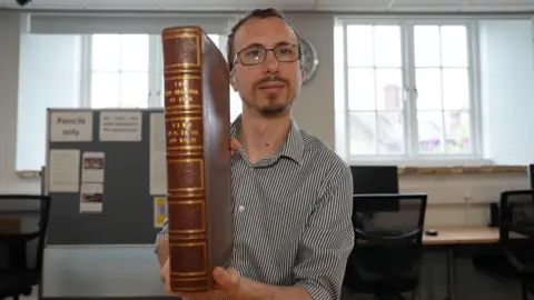 Luke Shackell sitting down at a table. He is looking directly at the camera and smiling. He is wearing a black and white striped shirt and has short dark hair and facial hair and is wearing a pair of glasses. He is holding the Red Register up to the camera.