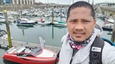 George is wearing a grey hoodie and a backpack and stands on a marina walkway, with rows of moored boats and yachts behind them. A bright red speedboat is tied up in the foreground, and apartment buildings and harbour infrastructure are visible in the distance under an overcast sky.