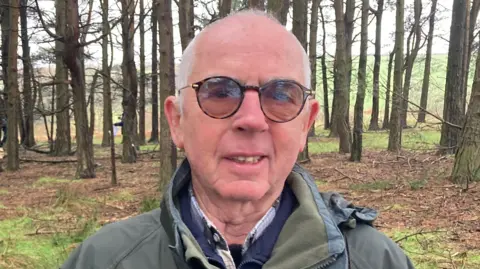 Carla Fowler/BBC A man with very short white hair wearing dark framed tinted glasses and a khaki green waterproof coat. He is standing outdoors, with a forest behind him and a view of moorland behind the trees.