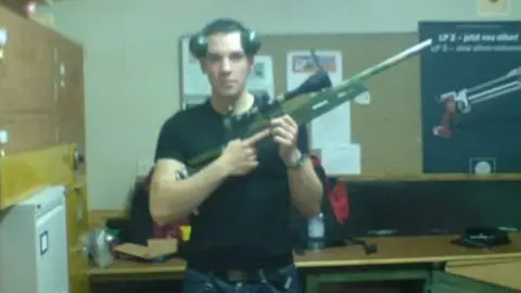 Finlay MacDonald in a room holding a gun. He is wearing a black tshirt and blue jeans, and has ear protectors on top iof his head.