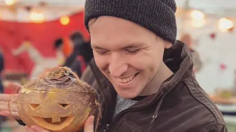 RYAN MORRISON Ryan Morrison is pictured holding a carved turnip and smiling. He is looking down. He is wearing a dark grey beanie hat and a brown jacket. The background behind him is blurred.