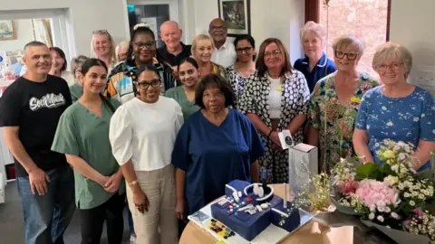 Fiona Callow/BBC A mixed group of people smile at the camera, some in work uniform, stand together, behind a table with a birthday cake, flowers and balloons on it.