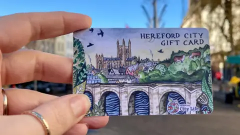 Hereford BID A woman's hand with gold rings holds up a small credit card-sized gift card with a drawing of the Hereford skyline and "Hereford City Gift Card" written in the top right corner.