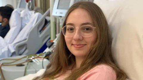 Sophie wearing a pink top and lying on a bed as stem cells are removed from her blood by a machine she is connected to. The Apheresis machine extracts the cells which she is donating to a stranger