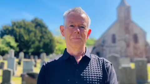 BBC Man - Charles Roberts-McIntosh - standing in graveyard, with Arbuthnott Church in background, under a blue sky.