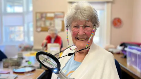 Nina Gerlack, one of the members, smiling at the camera in her arts and crafts class. She has a magnifying glass around her neck and is wearing glasses with a bead necklace attached to them. On the table are arts and crafts equipment.