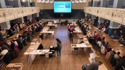 Oxford City Council election count in Oxford Town Hall. There are tables full of ballot papers that people are counting through.