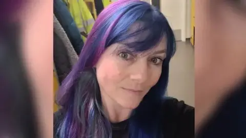 Family handout Rebecca Ashmead, with purple toned hair and a fringe. She is looking at the camera in a selfie-style shot. Behind her there are some jackets, including a high-vis coat and a door. She is wearing a dark jumper.