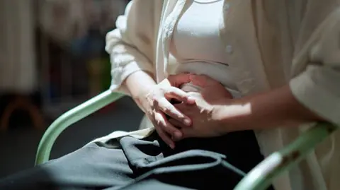 Getty Images Stock image of a woman wearing a white shirt, white vest and black trousers, sitting in a green chair. She's holding her hands over her lower stomach as if in pain. 