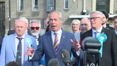 Farage in a blue suit and striped purple tie flanked by Reform councillors.
