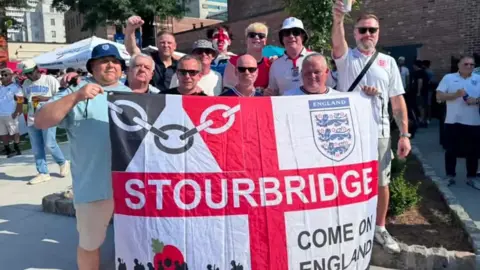 A group of men in football shorts, many in sunglasses and bucket hats, stand in a public square holding a flag featuring the St George's cross. The quarters of the flag are filled with the England football crest, Black Country flag, Help for Heroes logo and "Come on England". 