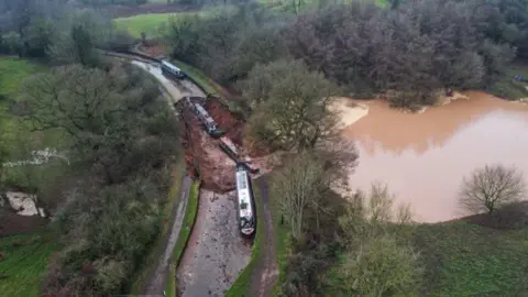 PA Media An aerial view of a grass-edged canal with a number of narrowboats in it and two at the bottom of a large muddy hole, with a large brown body of water to the right hand side