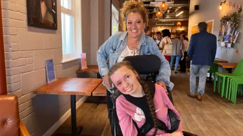 A woman smiling at the camera wearing a denim jacket and white top. She is standing behind a woman in her twenties who is sat at a slight angle in a wheel chair. She is wearing a pink Nike top and smiling at the camera.