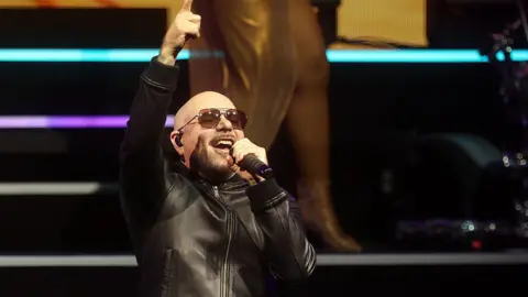 Pitbull, a bald man wearing aviator sunglasses, singing on stage. He is wearing a black leather jacket.