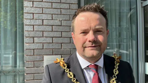A man with short dark hair standing in front of a building. He wears a black suit, white shirt and red tie with a blue stripe on it. There is a gold chain hanging from his shoulders to represent his role as a mayor. 
