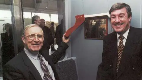 Nexus Tony Ridley inside a Metro Train with another man. Both have grey hair and are dressed in dark suits. They are both smiling.