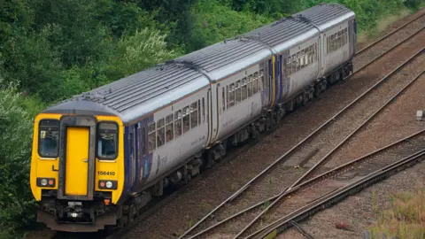 A Northern rail train on a track backed by green shrubs and bushes