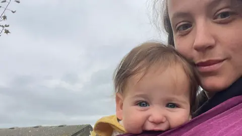 Hannah Howell Mother Hannah Howell (right) has brown hair and blue eyes and looks into the camera. While her 7-month-old baby with blue eyes and brown hair strapped to her chest smiles into the camera. 