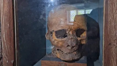 A mummified skull sitting on a small wooden block behind a glass screen.  
