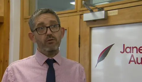 A middle aged man with greying hair is wearing dark framed glasses, a pink and white pinstripe shirt and a dark blue tie as he faces the camera. He is standing in front of wood and glass doors in a hallway.