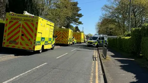 Three ambulances and a police van are parked along a residential road.