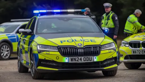 A black, yellow and blue Skoda police car with its blue lights on leaves a car park where two other police cars and three police officers are. 
